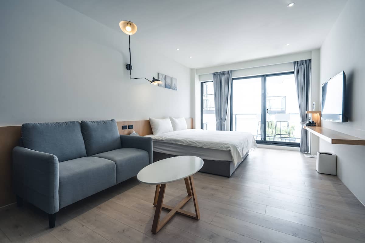 Una habitación de hotel moderna con un sofá gris, una mesa redonda blanca, una cama con ropa de cama blanca, un ventanal con cortinas y un televisor de pantalla plana en la pared.