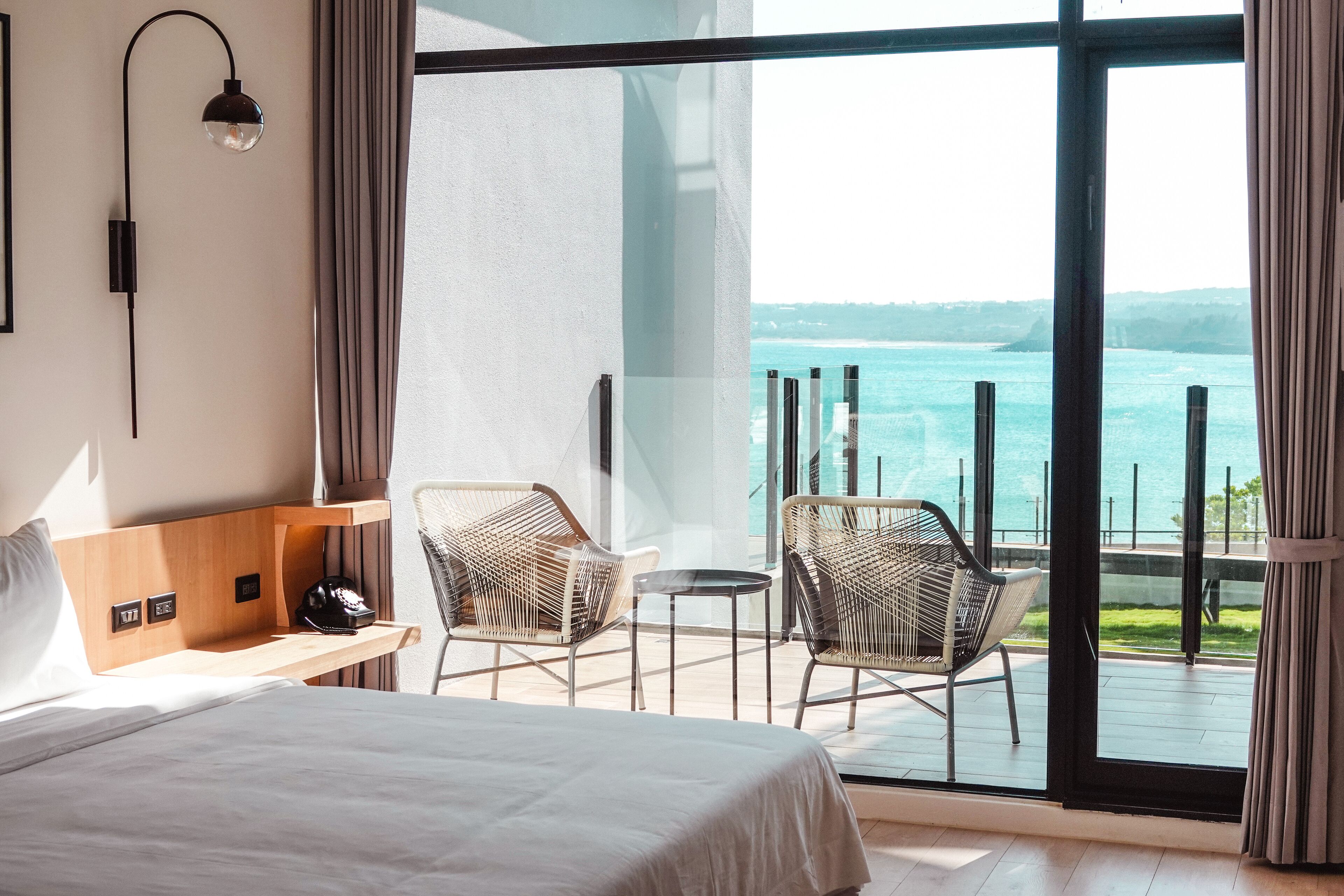 Une chambre d’hôtel avec un grand lit, deux fauteuils en osier, une petite table et une vue sur la mer par de grandes fenêtres.