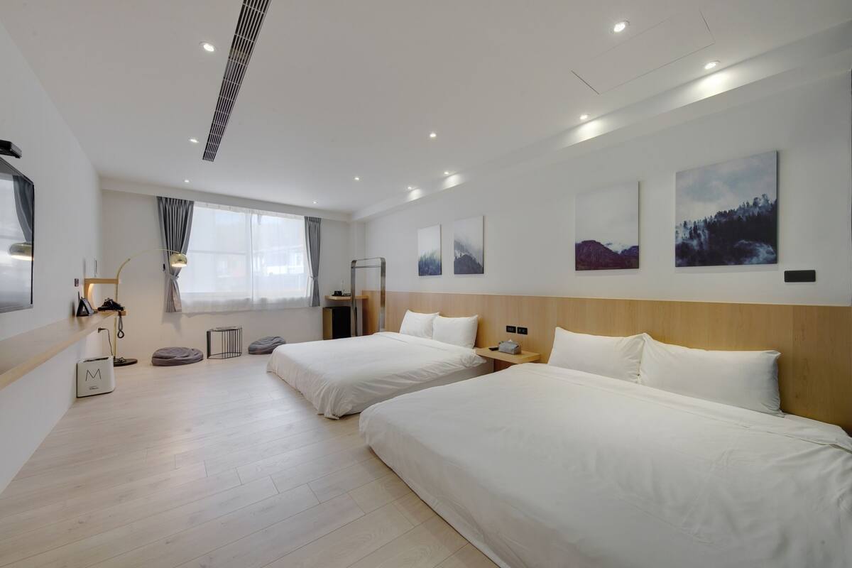 Habitación de hotel con dos camas, un ventanal con cortinas, un escritorio con lámpara y una obra de arte en la pared.