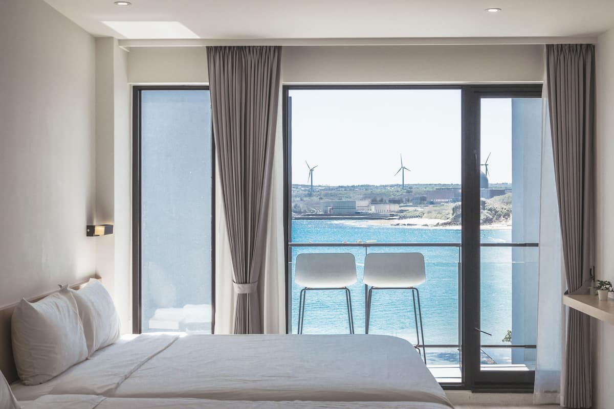 Una habitación de hotel moderna con una cama grande, dos sillas junto a un balcón con vista al mar y un paisaje urbano al fondo.