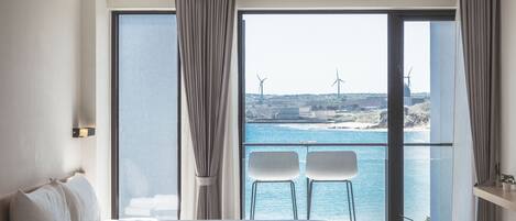 A modern hotel room with a large bed, two chairs by a balcony with a sea view, and a cityscape in the background.