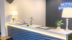 En moderne receptionsdisk på et hotel, med en taktekstureret blå væg, et ur og et neonskilt, der viser "HOTEL L'IMPERIA".