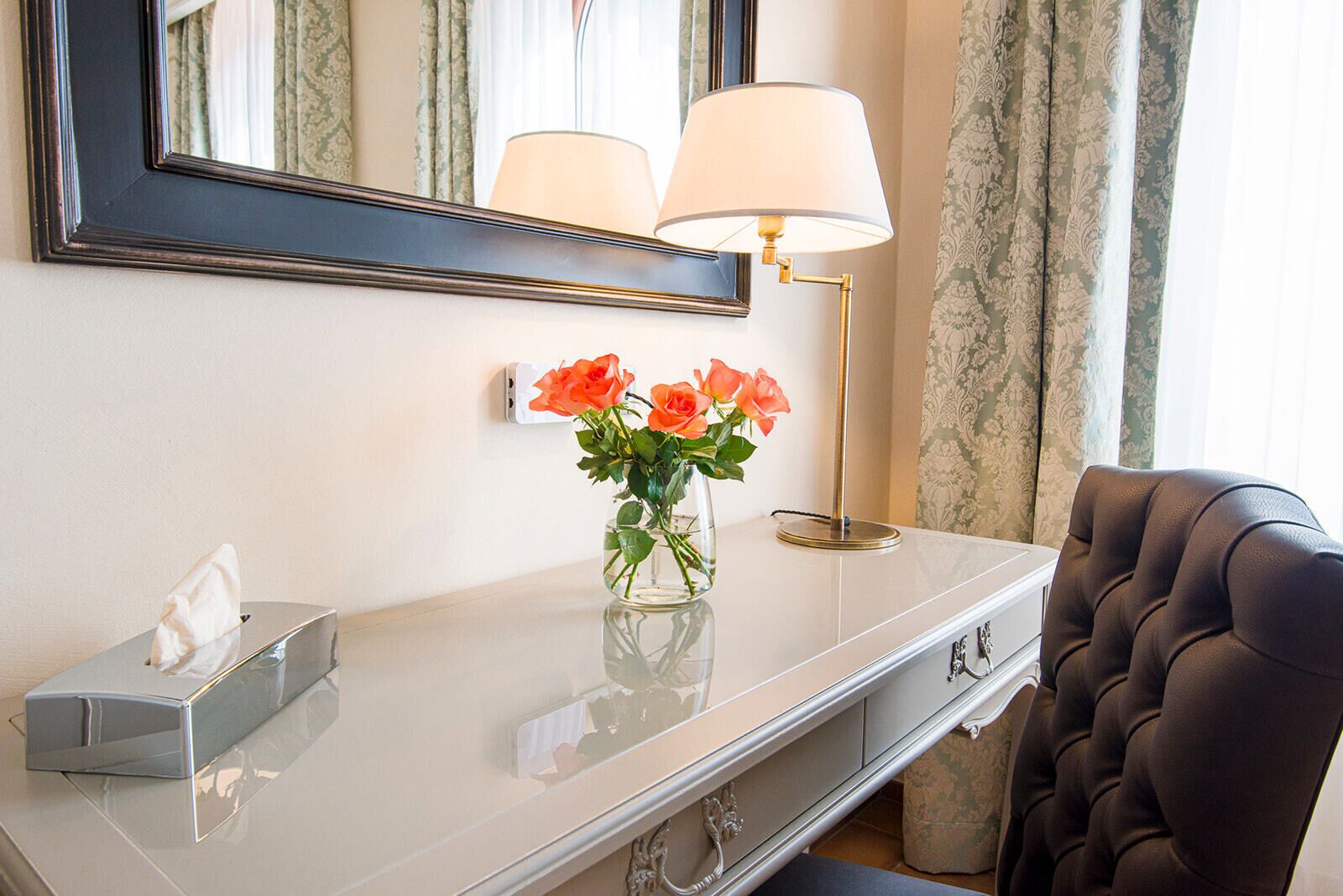 Ein Hotelzimmer mit Schminktisch, Spiegel, Lampe, einer Vase mit Blumen und einem Sessel.
