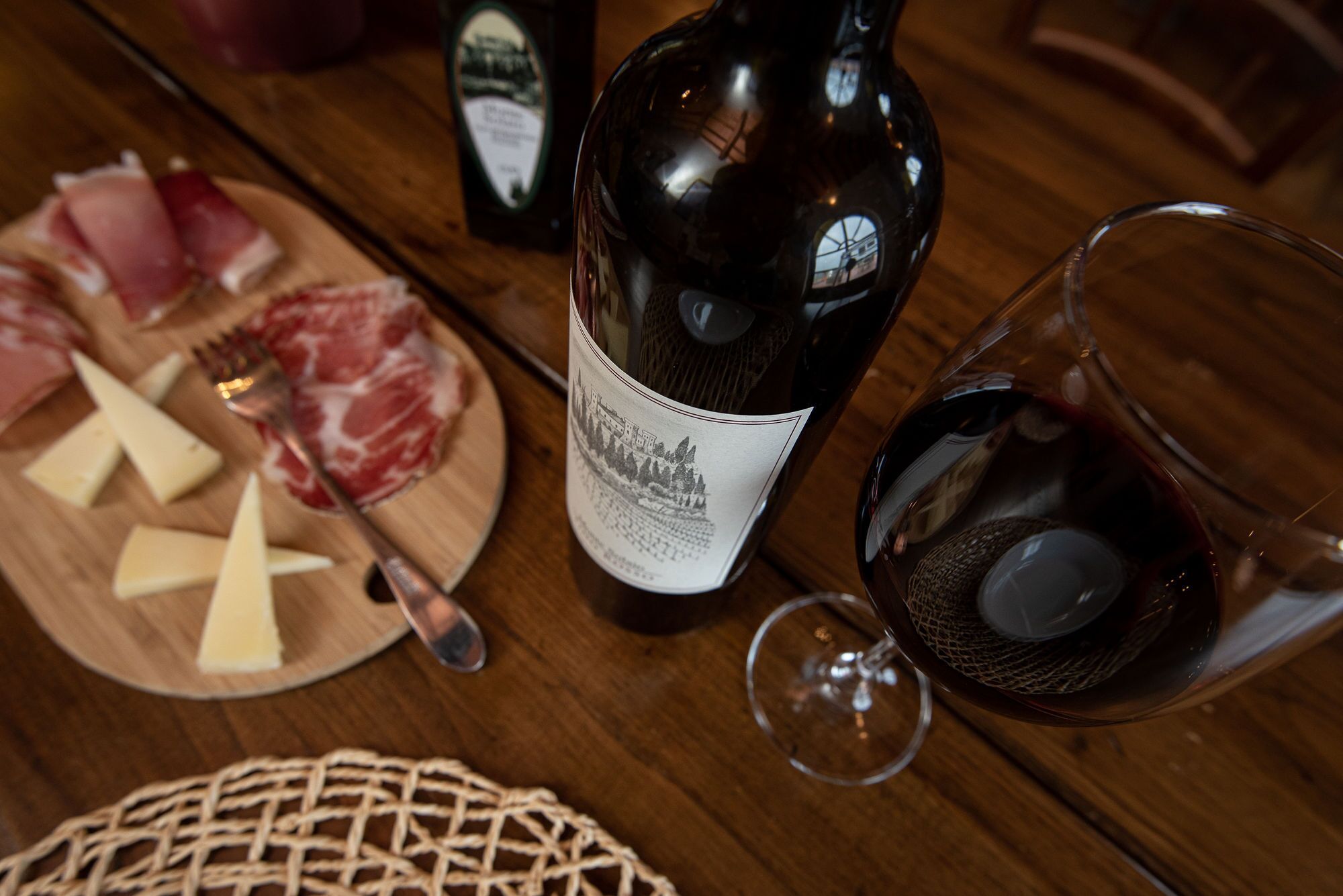 Una mesa de madera con una botella de vino, una copa de vino tinto, queso y embutidos.