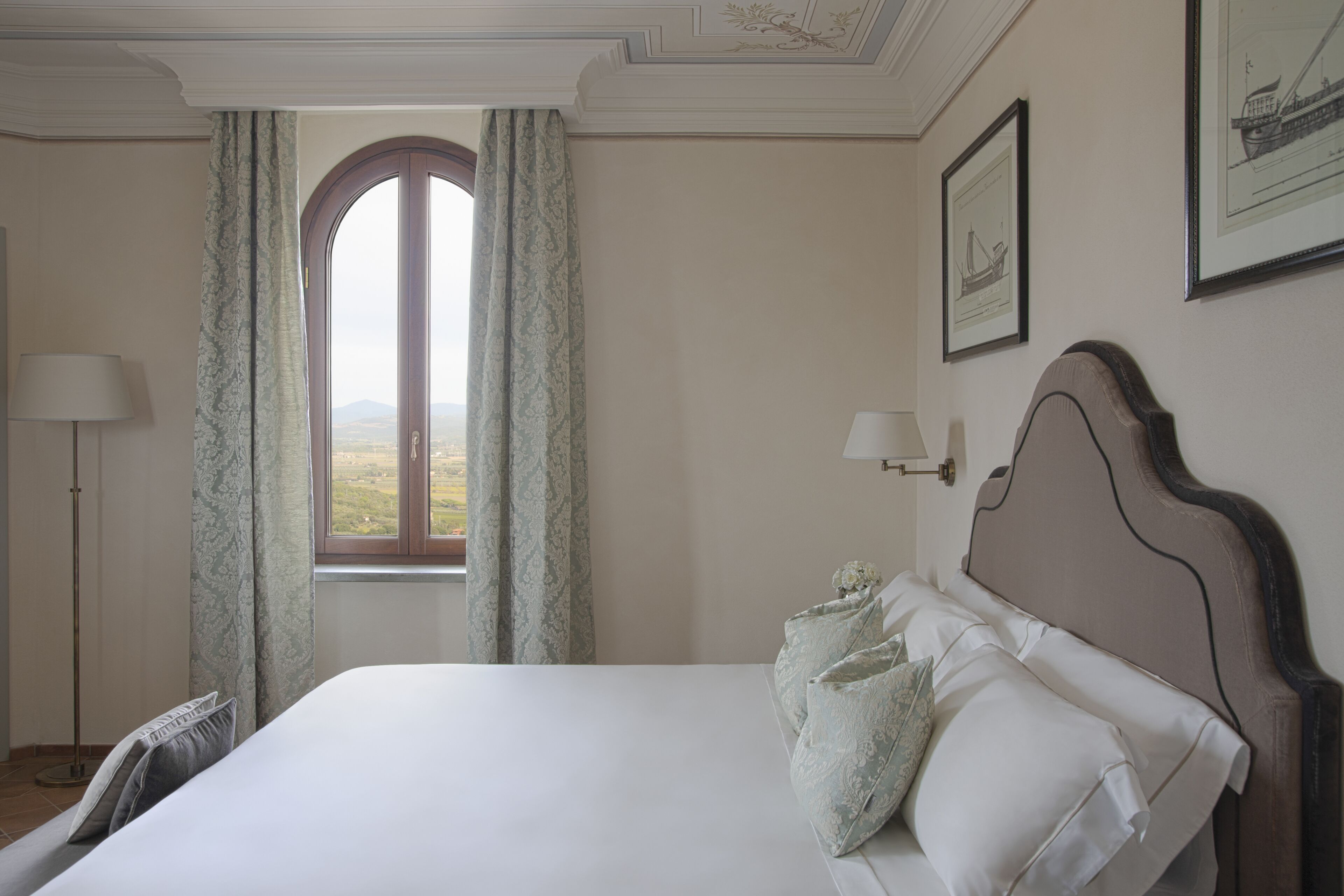 A bedroom with a large bed, a window with curtains, a bedside table with a lamp, and framed artwork on the wall.
