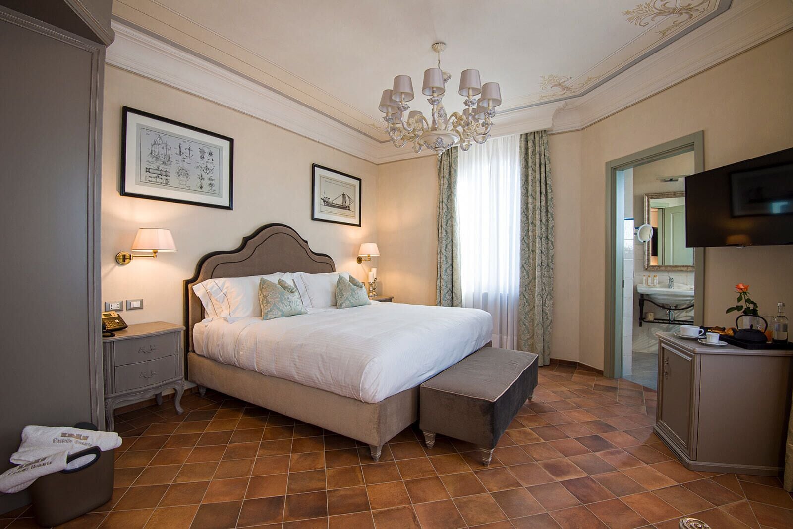 A well-appointed hotel room with a large bed, a bedside table with a lamp, a bench at the foot of the bed, a television, and a bathroom visible through an open door.