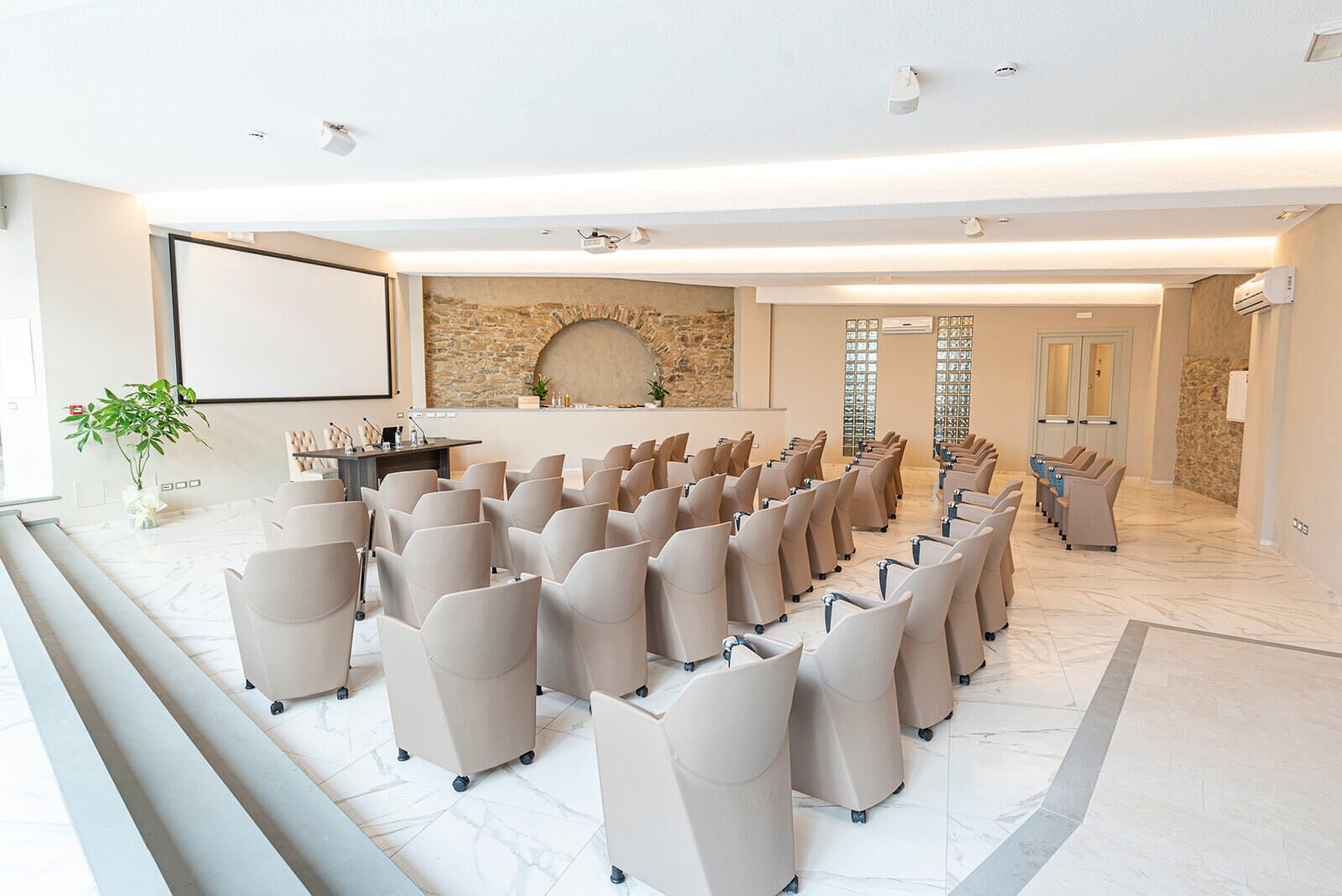 Uma sala de conferências com fileiras de cadeiras, uma tela grande, uma parede de pedra e uma área de recepção com balcão e cadeiras.