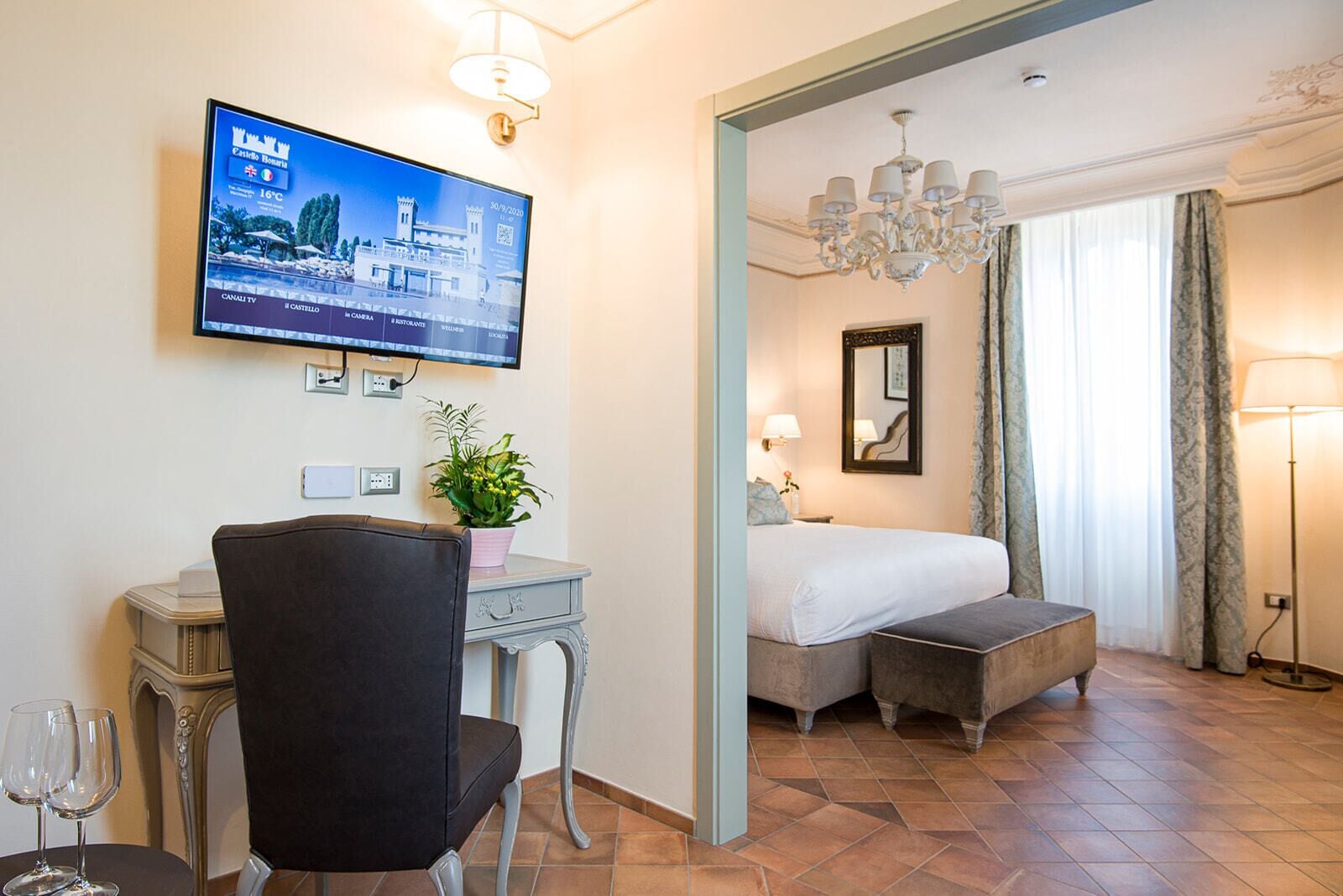 Una camera d'hotel con un letto, una scrivania con una sedia, una TV a parete e una pianta in vaso.
