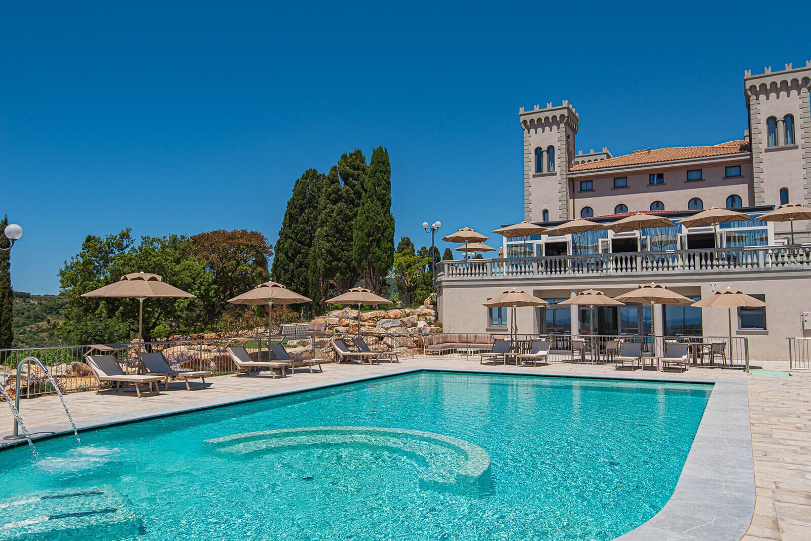 Un espace piscine avec des chaises longues et des parasols, un grand bâtiment doté d’une tour, et un ciel d’un bleu limpide.
