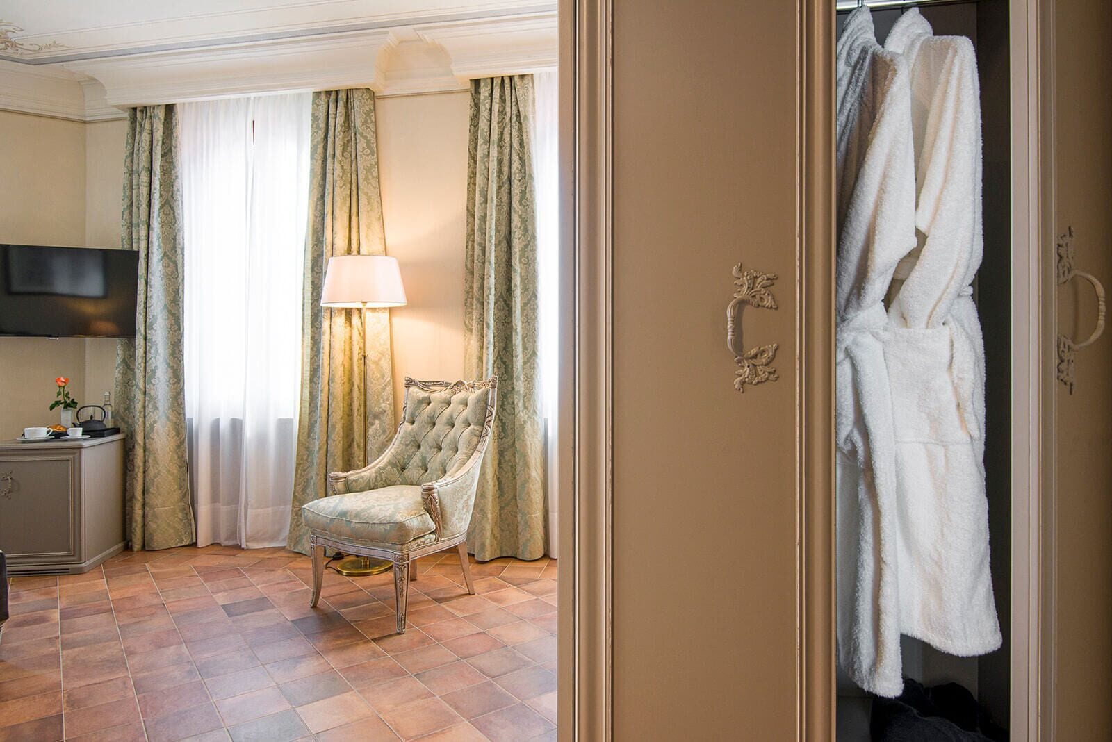 Quarto de hotel com poltrona estofada, mesa de cabeceira com abajur e guarda-roupa com roupões.