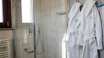 Un área de ducha con puerta de vidrio, un cabezal de ducha manual y un perchero con dos batas de baño blancas.