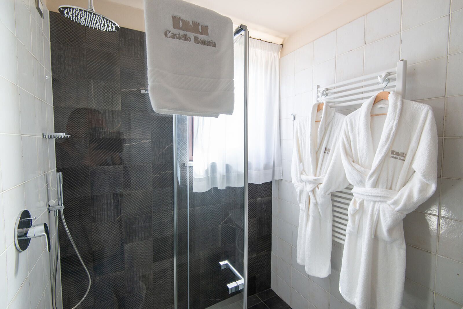 A hotel bathroom with a glass shower enclosure, a towel rack with a white towel, and a robe hanging on the wall.