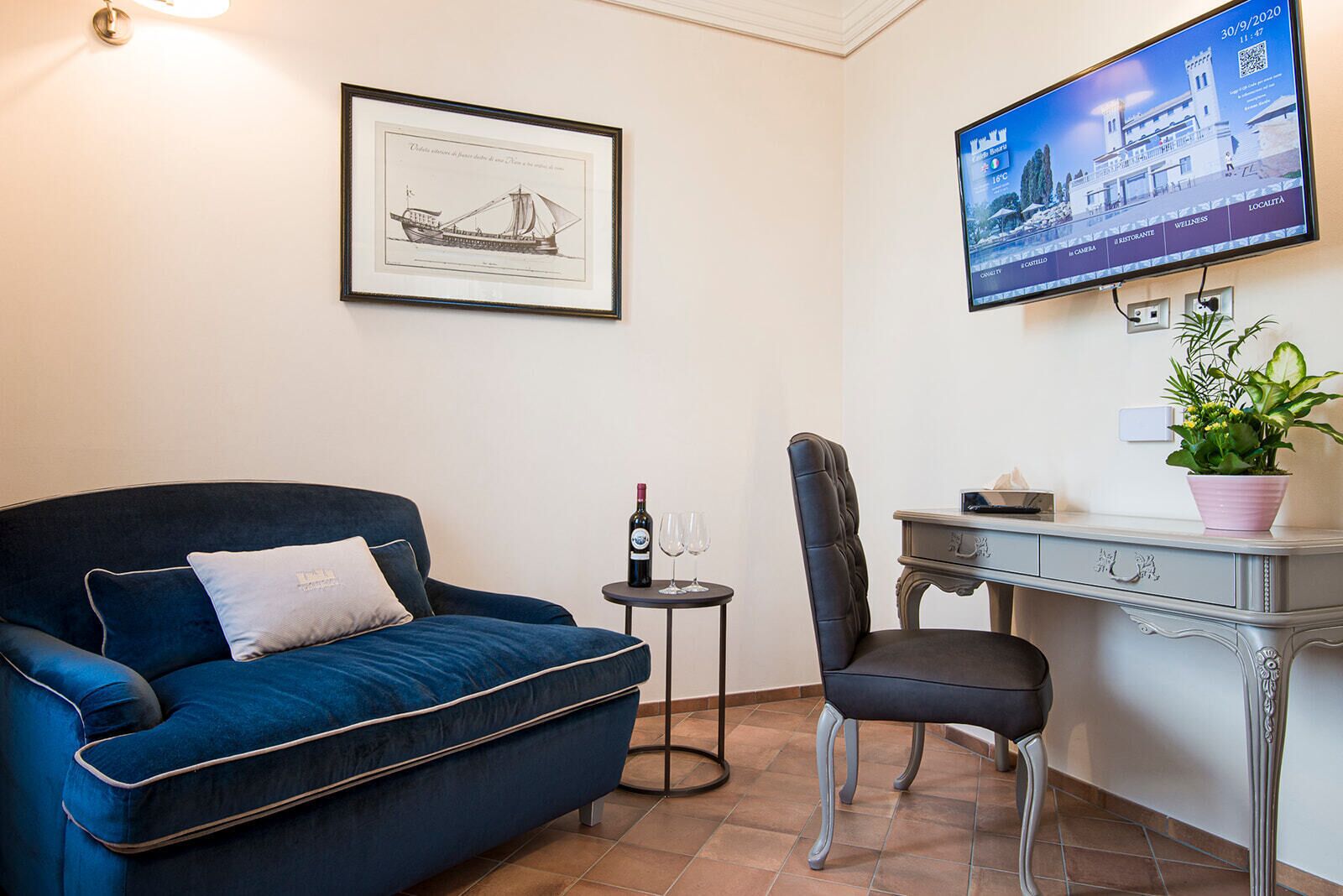 A room with a blue sofa, a desk, a chair, a small table with a wine bottle and glasses, a framed picture, and a TV on the wall.