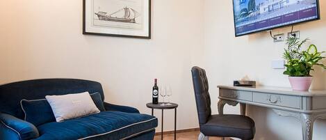 A room with a blue sofa, a desk, a chair, a small table with a wine bottle and glasses, a framed picture, and a TV on the wall.