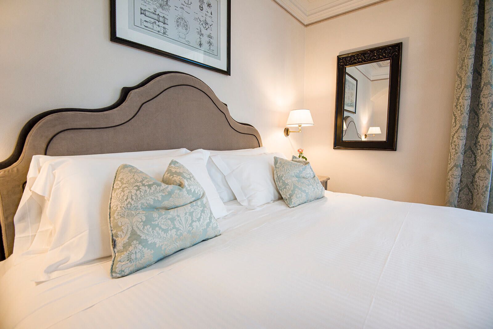 A neatly made bed with white linens and decorative pillows, a wall-mounted mirror, and a framed picture above the bed.