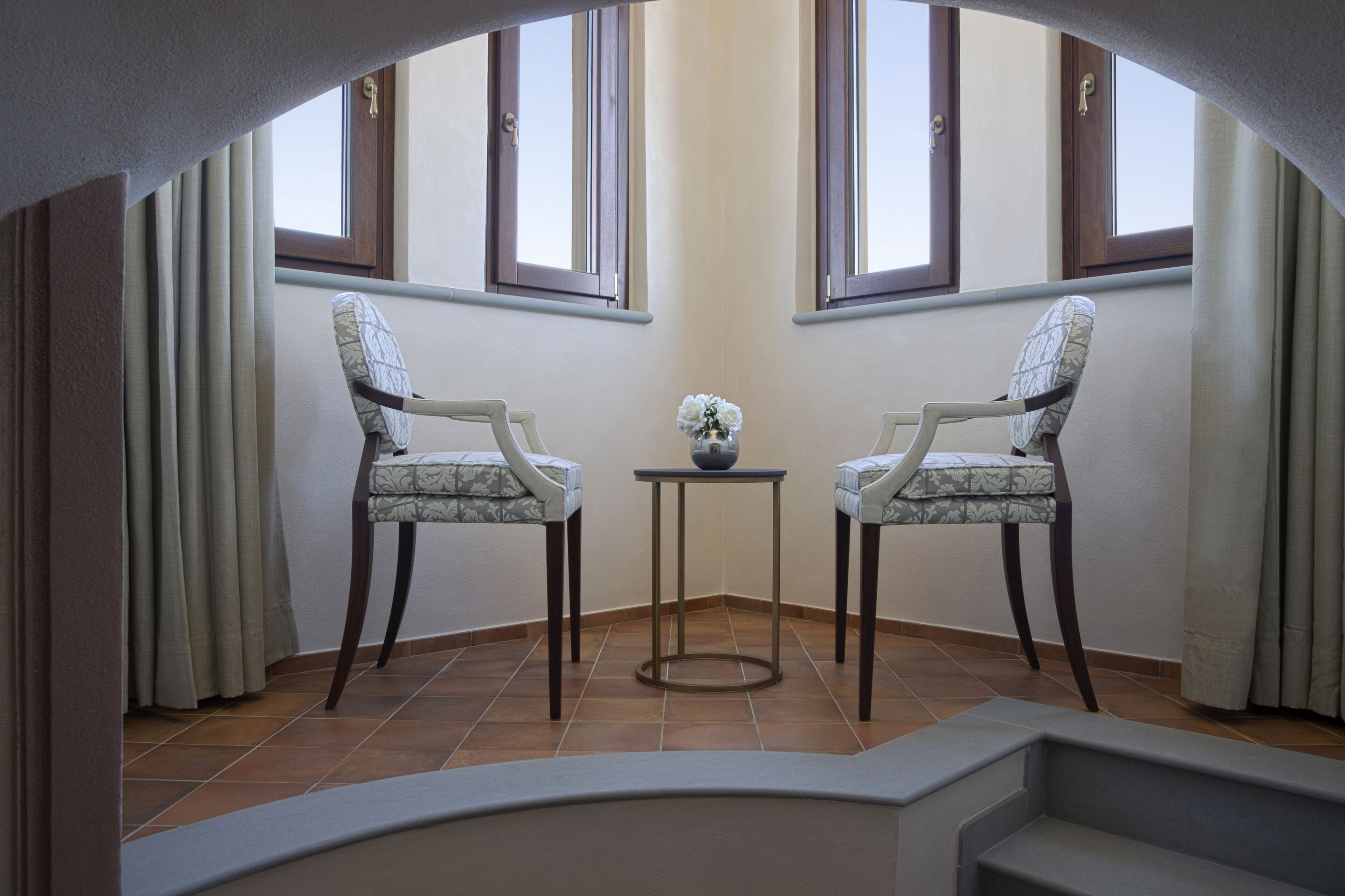 Una habitación con dos sillas frente a frente, una mesita con un florero y vistas al exterior a través de una ventana con cortinas.