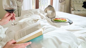 A person reading a book on a bed with a wine glass, a plate of food, and a bowl on the bed. - Castello Bonaria Wine & Spa Resort (Campiglia Marittima)