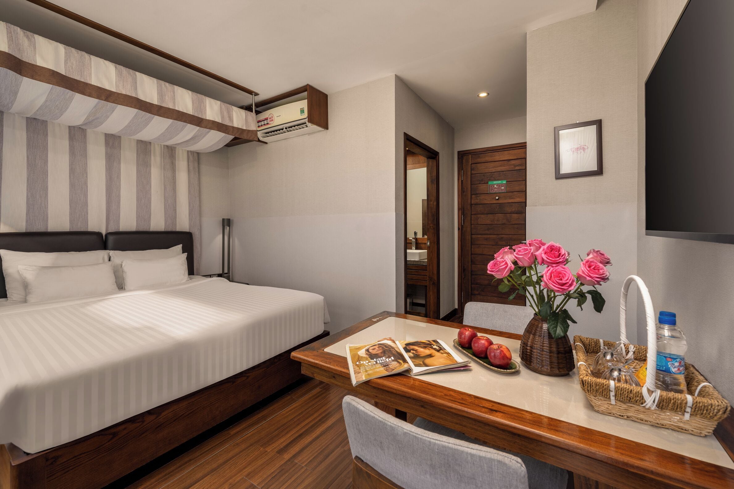 A hotel room with a bed, a wooden desk with a magazine, a vase with flowers, and a water bottle.