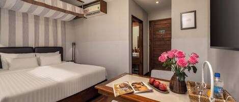 A hotel room with a bed, a wooden desk with a magazine, a vase with flowers, and a water bottle.