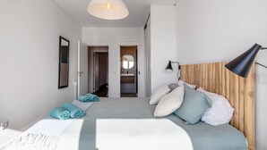 Quarto moderno com cama, luminária de cabeceira, janela grande e vista para um banheiro.