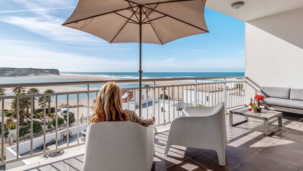 Uma varanda com um grande guarda-sol, cadeiras brancas, uma mesa pequena com flores e vista para a praia e o oceano.