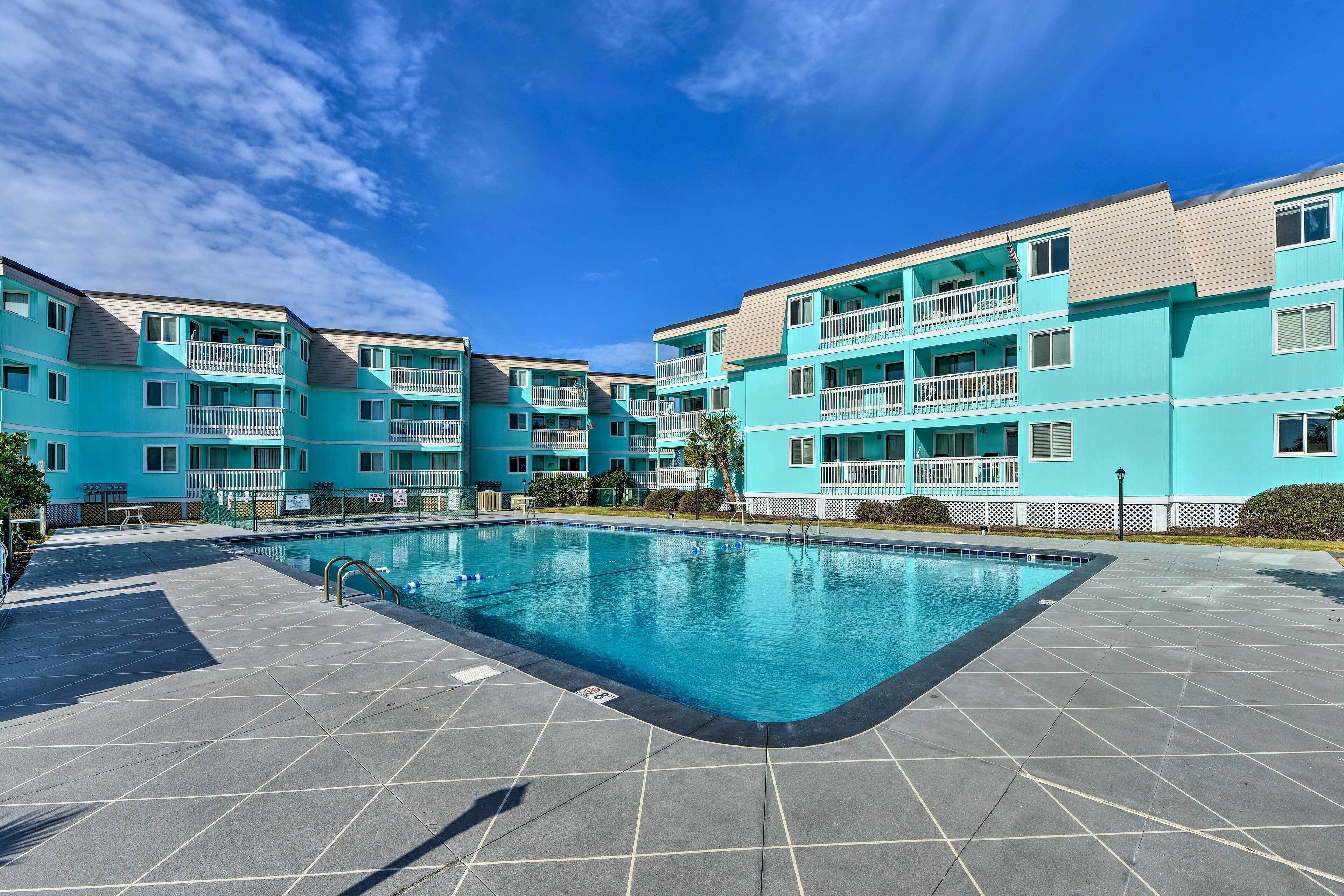 Un immeuble turquoise à plusieurs étages, doté d’une piscine, d’un espace extérieur aménagé et surplombant un ciel d’un bleu limpide.