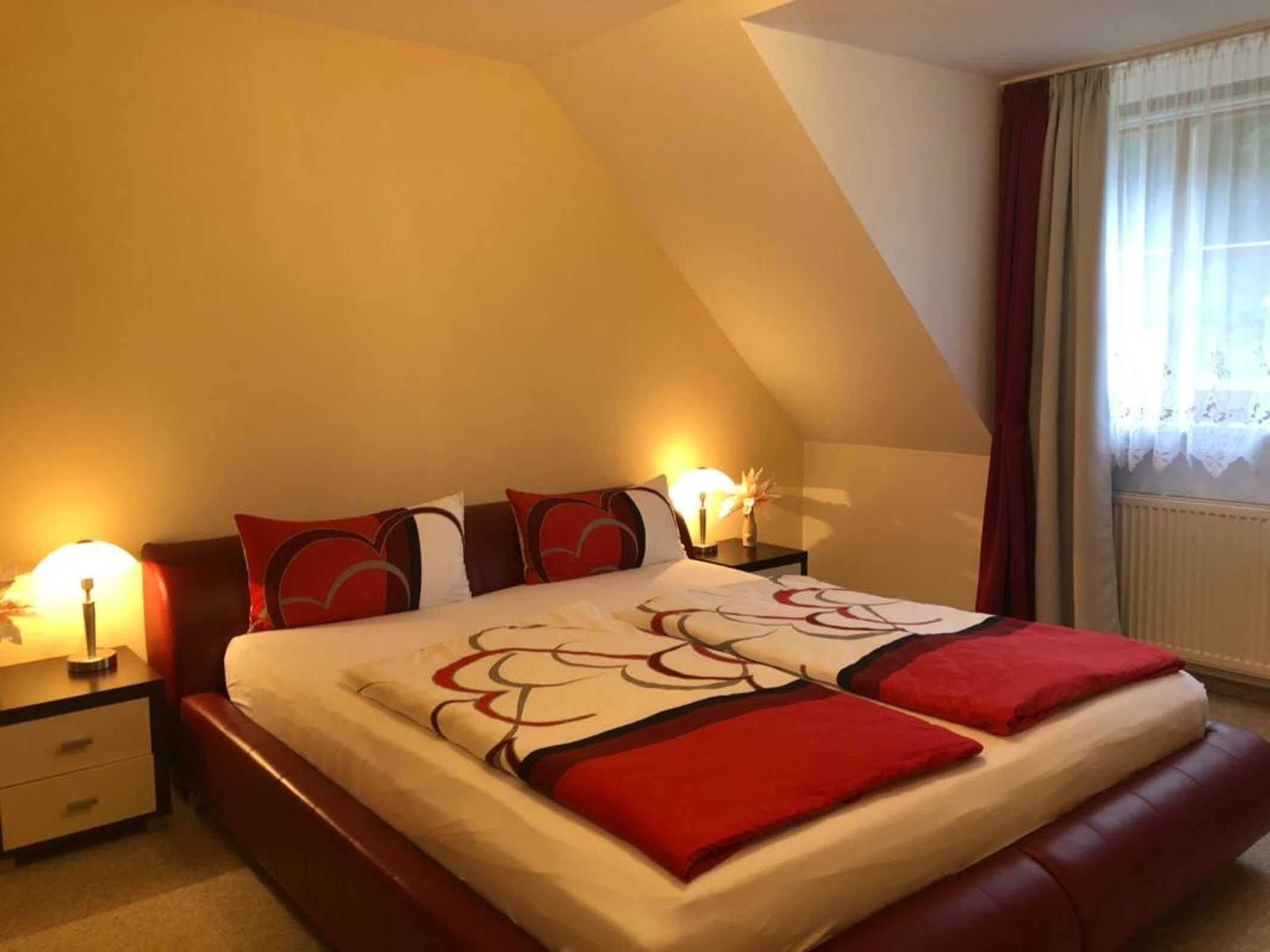 A bedroom with a sloped ceiling, a bed with red and white bedding, two bedside tables with lamps, and a window with curtains.