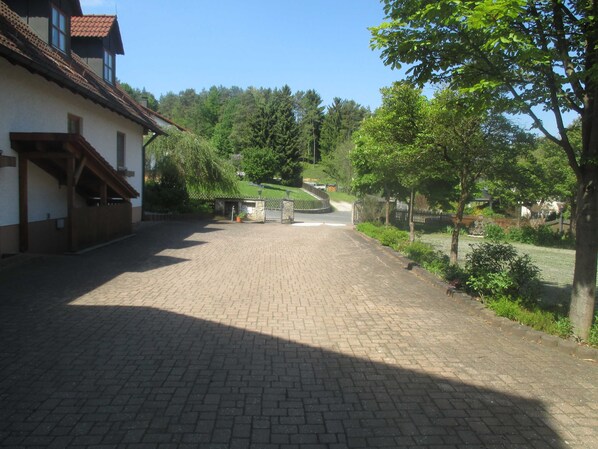 Une cour dallée avec un bâtiment sur la gauche, un petit portail et des arbres sur la droite.