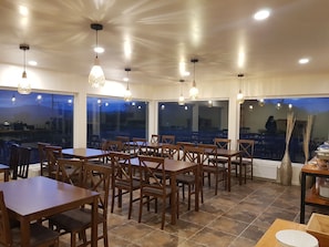 A restaurant with wooden tables and chairs, large windows, and hanging pendant lights.