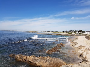 Ein Küstenstrand mit felsigem Ufer, blauem Himmel und weit entfernten Häusern.