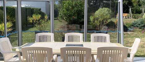 A covered patio with a table and chairs, surrounded by glass walls offering views of a garden and a house.