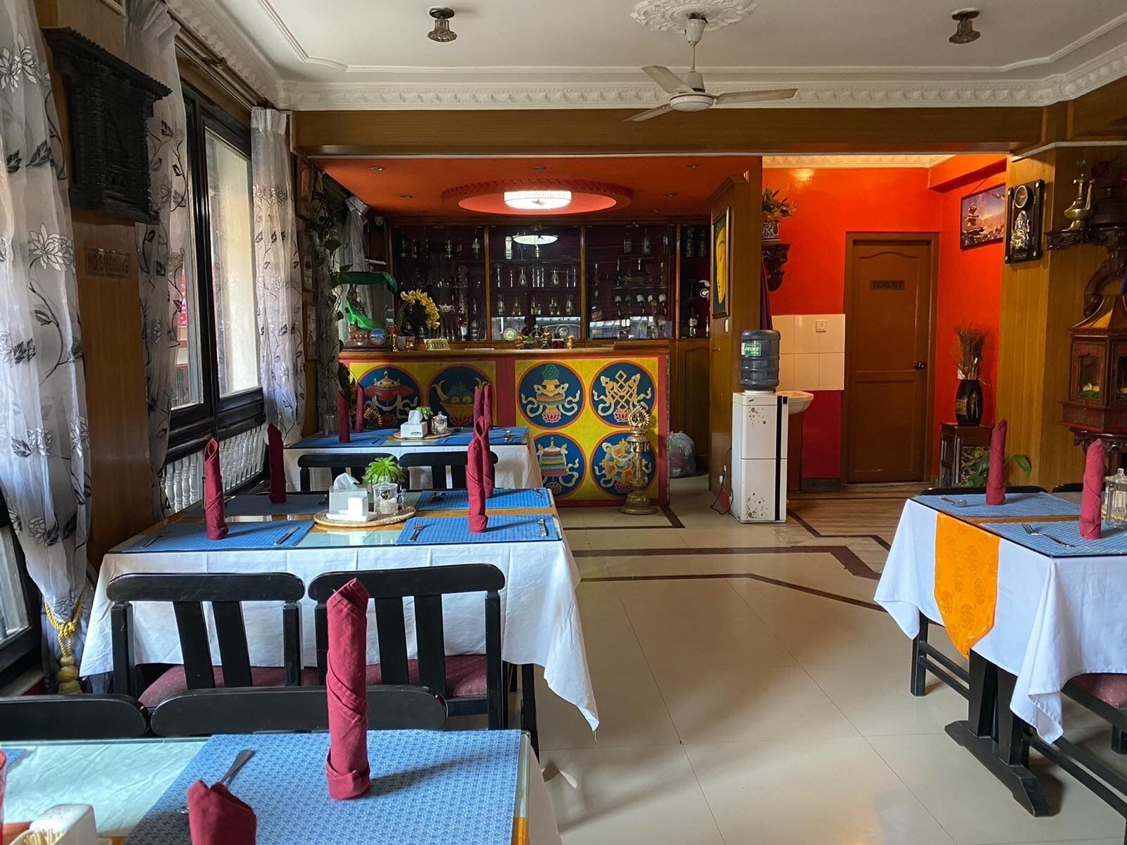 Un espace repas avec des tables préparées pour deux personnes, un bar agrémenté d’une fresque colorée et un distributeur d’eau.