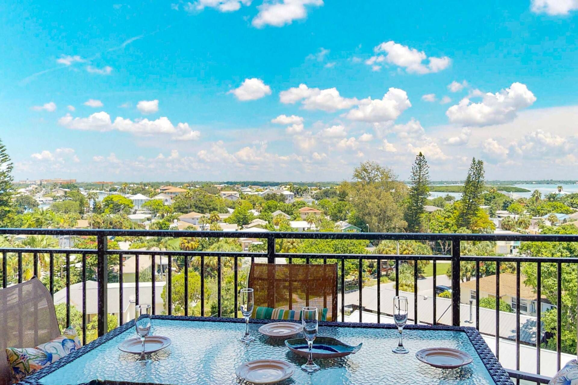 Un balcón con mesa de vidrio dispuesta para comer, con vistas a una zona residencial con casas y árboles.