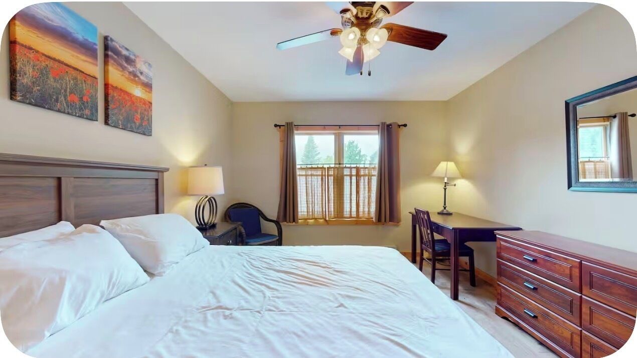 Une chambre à coucher comprenant un lit, un bureau, une chaise, une commode, un ventilateur de plafond et une fenêtre avec des rideaux.