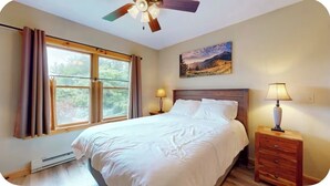 A bedroom with a bed, a ceiling fan, a nightstand with a lamp, a window with curtains, and a painting on the wall. - Quiet, Bright, Open, Rustic Next to Park and Pond with Mountain Views!  Apt 201 (Lake Placid)