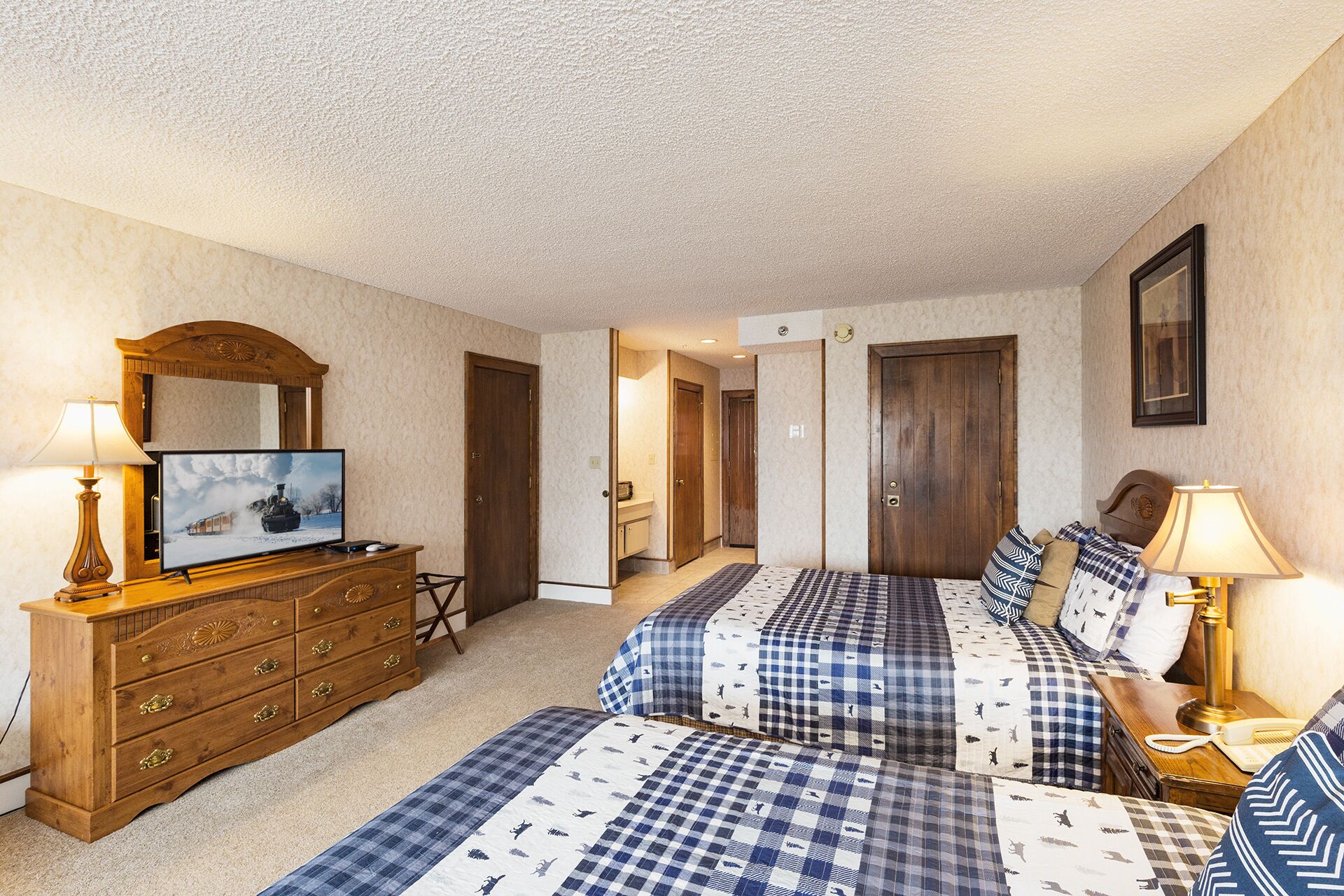 Quarto de hotel com duas camas, cômoda de madeira, televisão e escrivaninha com luminária.