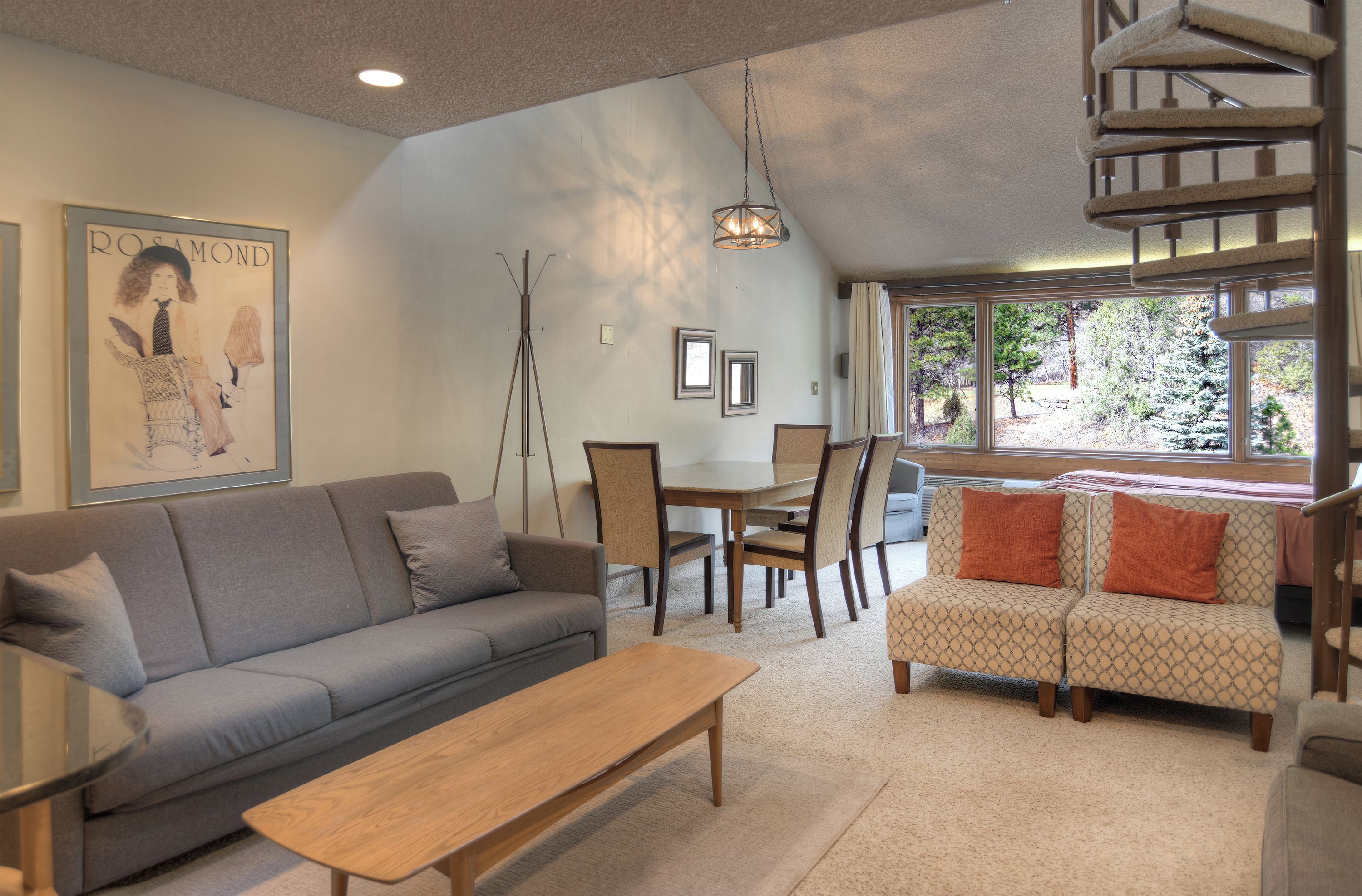 A living room with a gray sofa, a wooden coffee table, and patterned armchairs. A dining table with chairs is in the background.