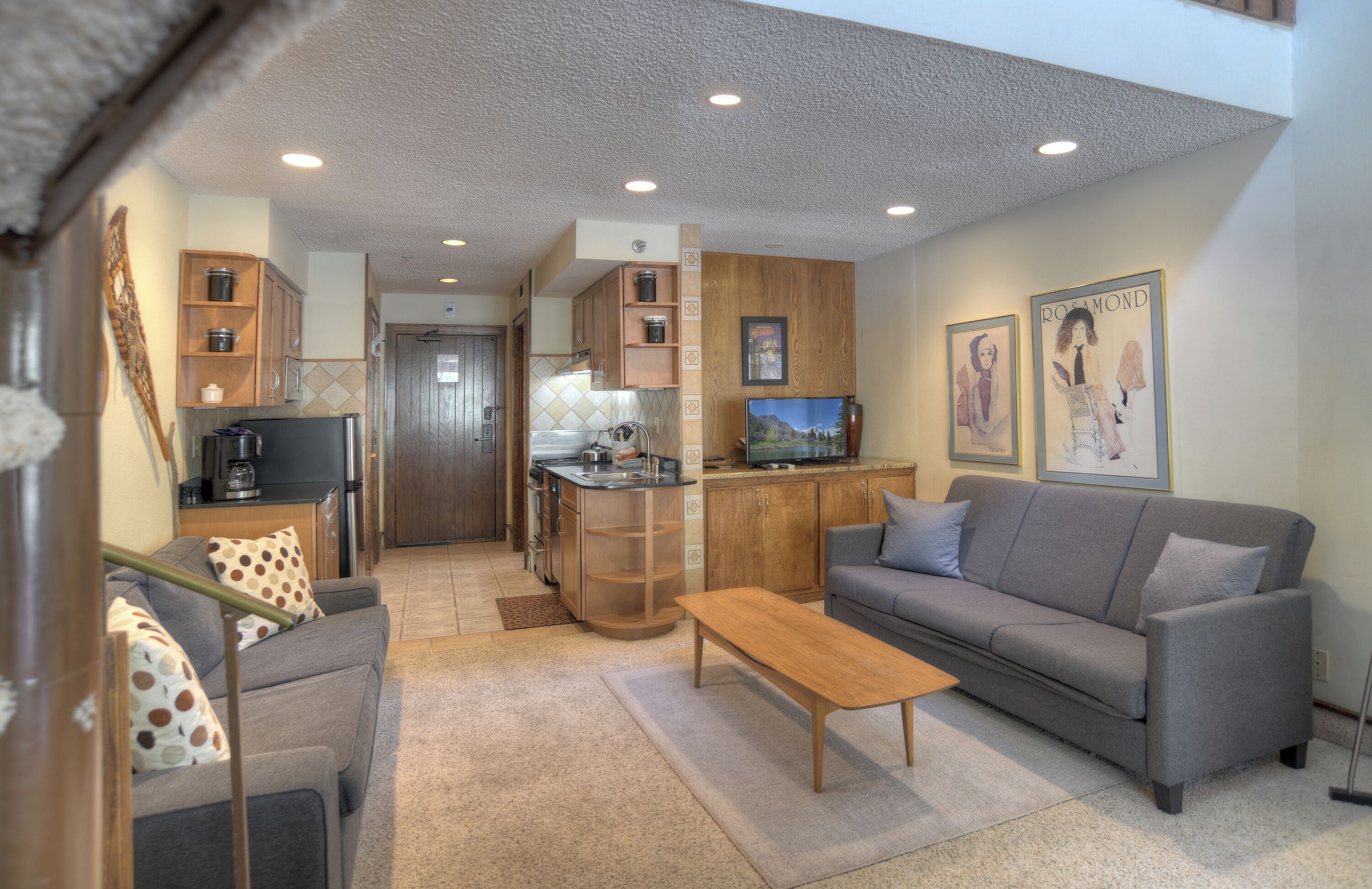 A compact living space with a kitchenette, grey sofa, wooden cabinets, and a television.