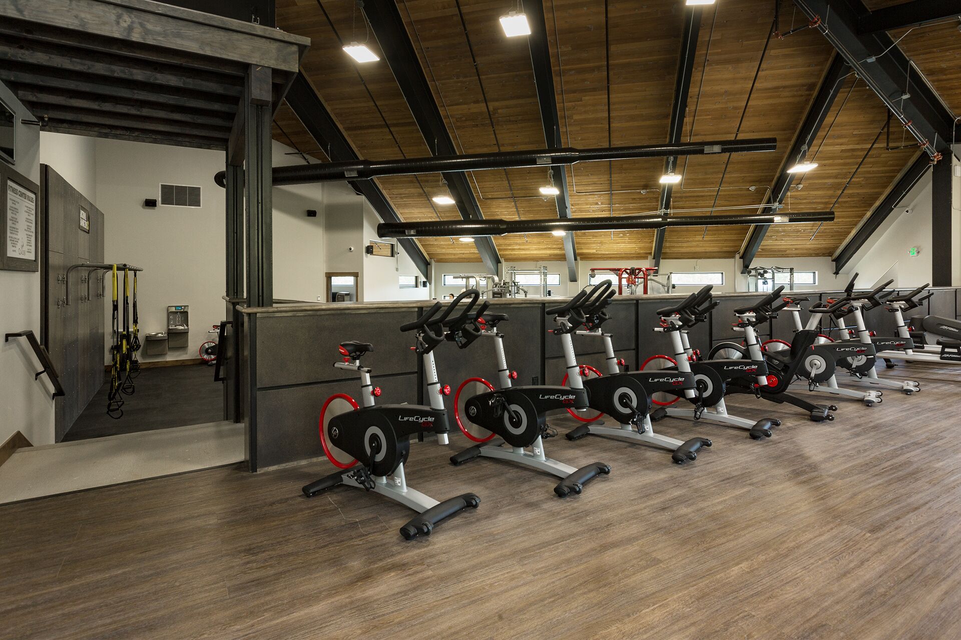 A gym with multiple stationary bikes, wooden flooring, and a high ceiling with exposed beams.