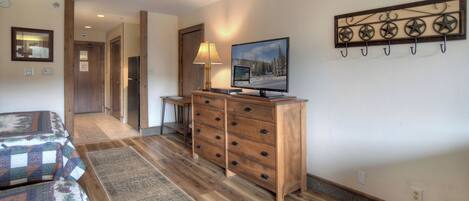 Une chambre d’hôtel avec une commode en bois, un téléviseur, un lit recouvert d’une couvre-lit à motifs et un couloir menant à une autre pièce.