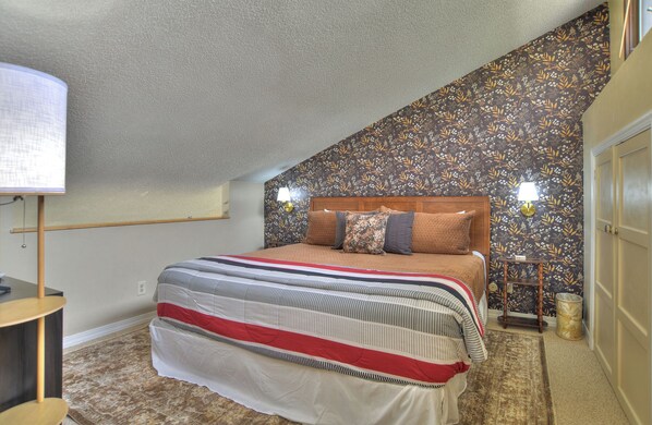 Un dormitorio con techo inclinado, una cama con ropa de cama a rayas, cabecera de madera y papel tapiz floral.