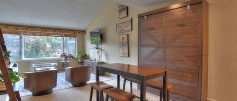 A room with a large wooden sliding door, a dining table with stools, a TV mounted on the wall, and a view of trees through the windows.
