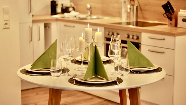 Una mesa de comedor dispuesta con servilletas verdes, platos y copas de vino, con velas y una máquina de café al fondo.