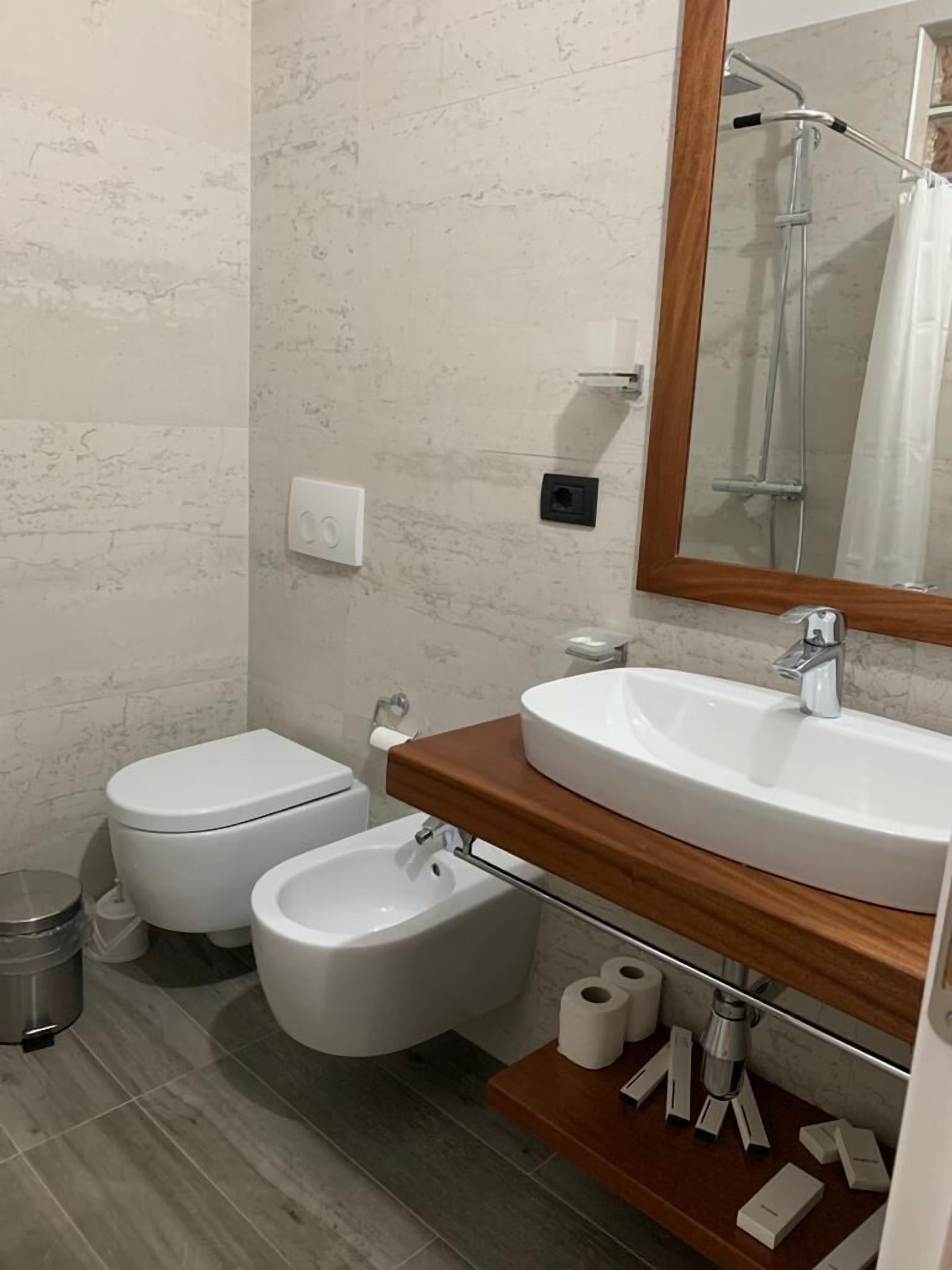 A bathroom with a toilet, bidet, sink, mirror, and a trash bin.