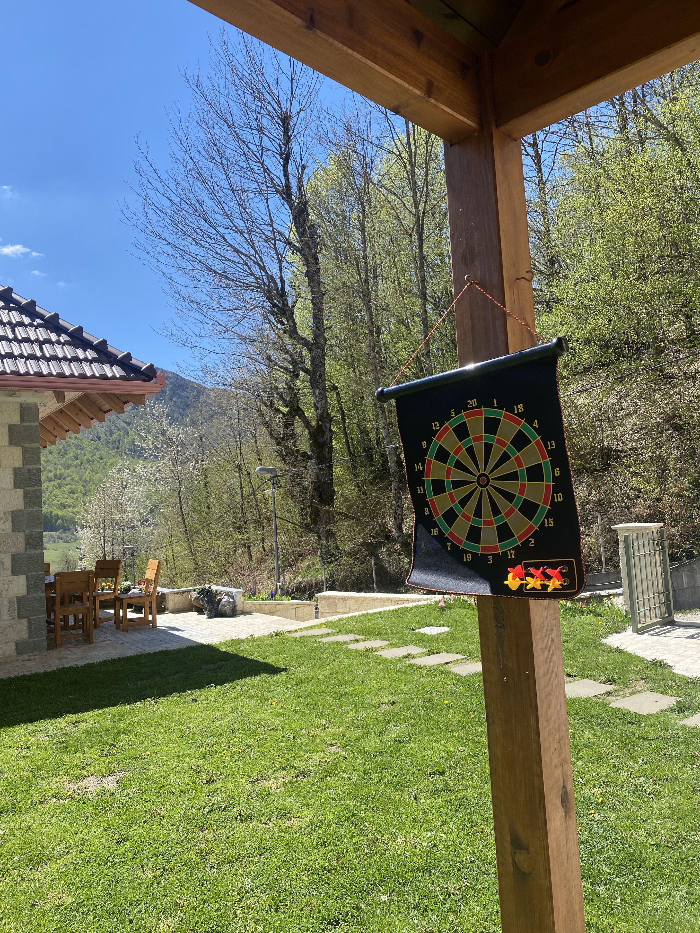 A dartboard mounted on a wooden post in a backyard with a stone patio, a wooden table and chairs, and a mountain backdrop.
