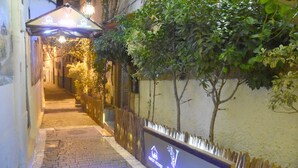 Uma passagem estreita com caminho de tijolos, cerca de madeira e placa indicando uma residência e restaurante.