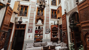 Decoração marroquina tradicional com portas de madeira elaboradas, uma área de estar central com almofadas e uma estante de livros.