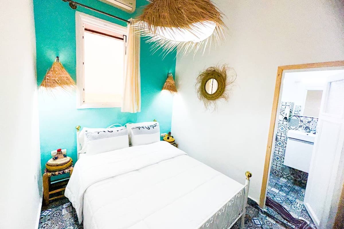 A room with a bed, a window, a wall-mounted air conditioner, a hanging light fixture, and a patterned floor.