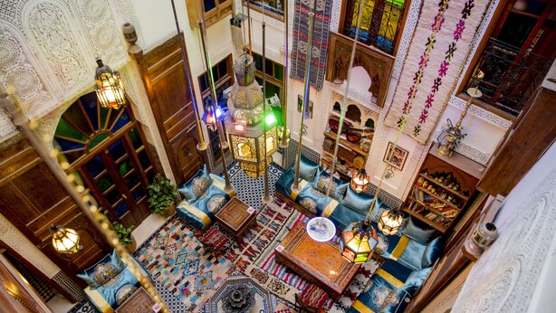 Um interior marroquino tradicional com decoração elaborada, um tapete estampado e uma área de estar central com almofadas e uma mesa de centro.