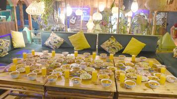 A dining area with a long table filled with various dishes and drinks, surrounded by a cozy seating arrangement with cushions and hanging lights.